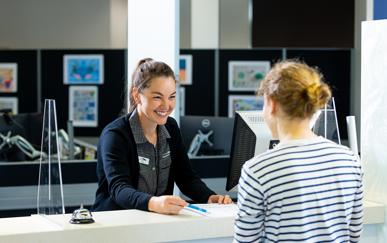 A smiling customer support team member leaning over a counting helping a customer in a striped top.