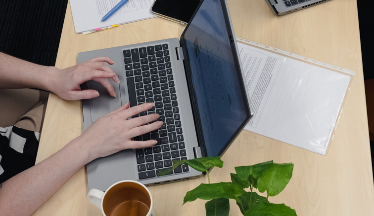 Close up of someone's hands typing on a laptop. A piece of paper, coffee cup and a plant on a light wooden desk.